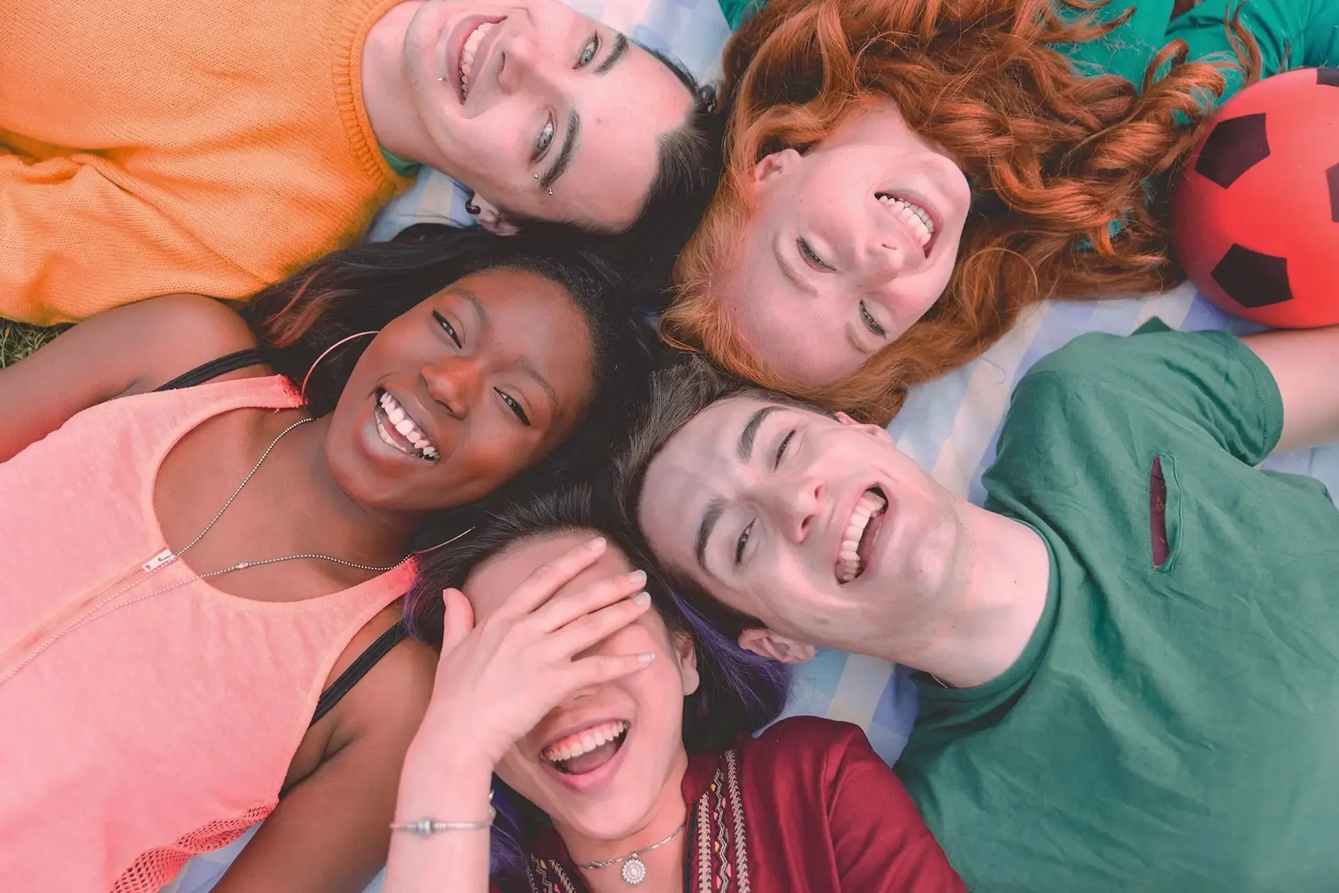 Smiling friends relax on a blanket in Twinsburg, OH; one hides their face after visiting Holt Orthodontics. Soccer ball nearby.