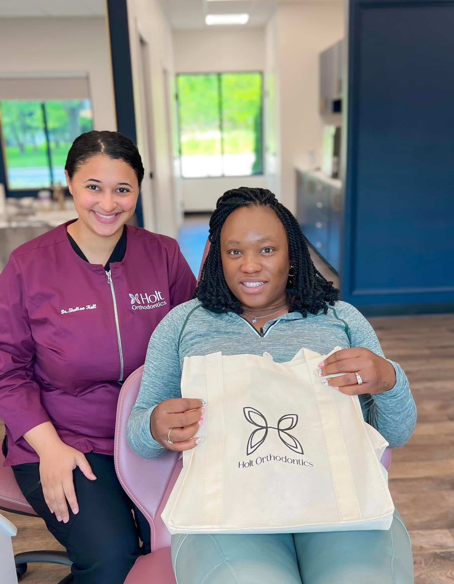Inside a modern clinic in Twinsburg, OH, a woman sits in a dental chair holding a Holt Orthodontics tote bag beside Dr. Shalise Holt in a maroon uniform.
