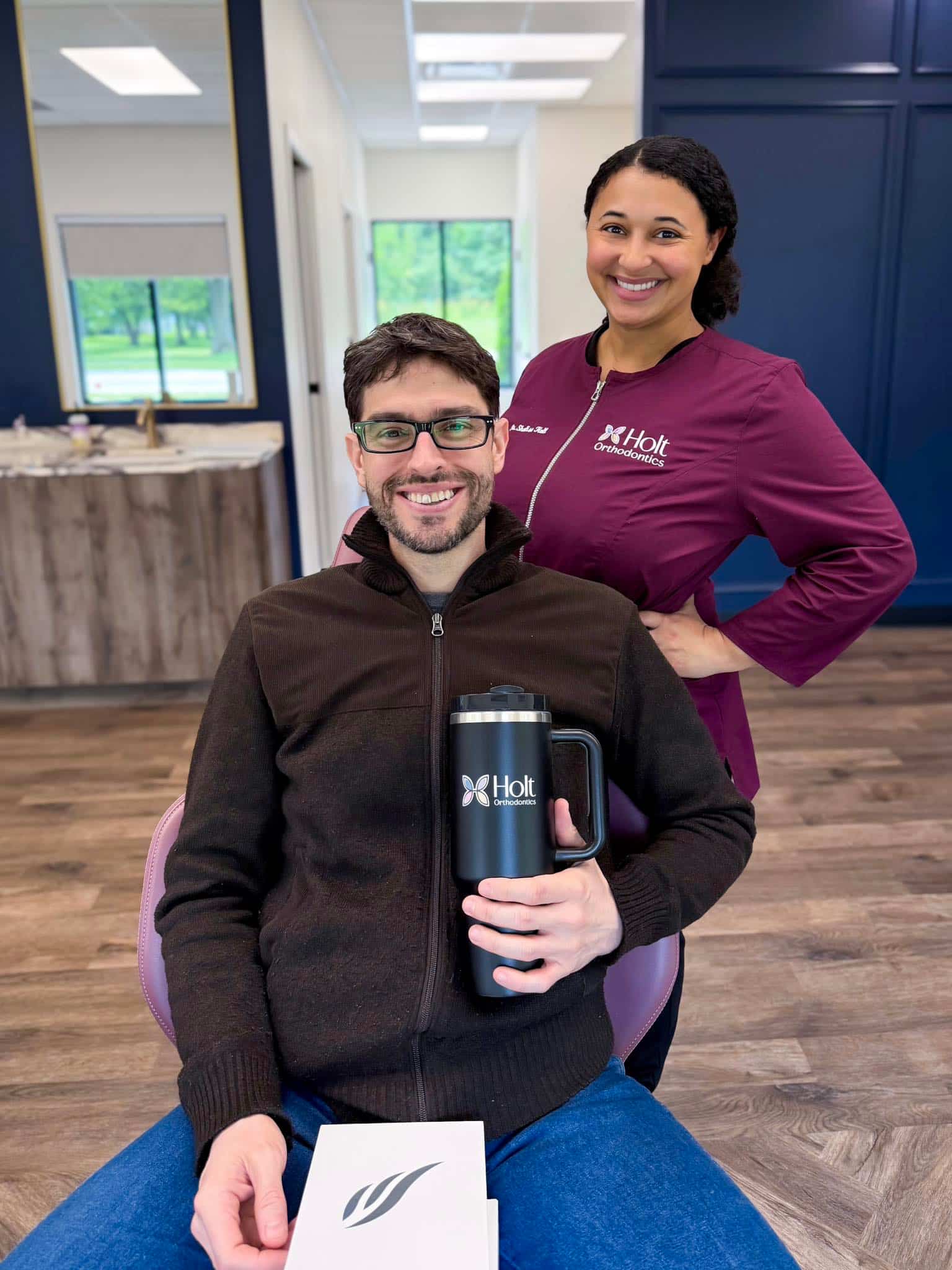 In an office with wooden floors and large windows, a man smiles with a mug and box while Dr. Shalise Holt of Holt Orthodontics in Twinsburg, OH, stands behind him in her dental uniform.