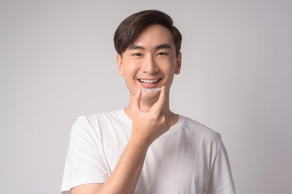 A young man smiles at the camera while holding an Invisalign aligner near his mouth, representing Holt Orthodontics in Twinsburg, OH.
