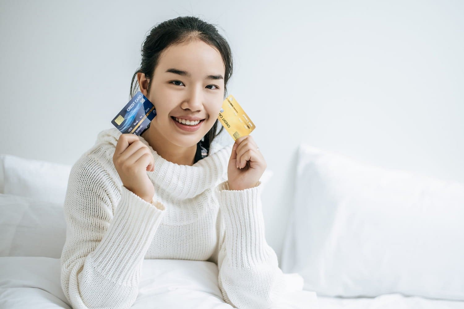 At Holt Orthodontics in Twinsburg, OH, a woman in a white sweater sits on her bed, smiling as she holds both a blue and a yellow credit card, ready to select the ideal braces payment plan.
