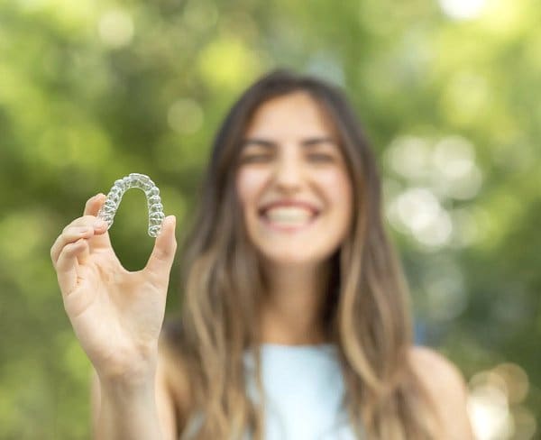 A woman smiles while holding a clear dental aligner in her outstretched hand, with a blurred green outdoor background, emphasizing that Invisalign at Holt Orthodontics in Twinsburg, OH may be covered by insurance.