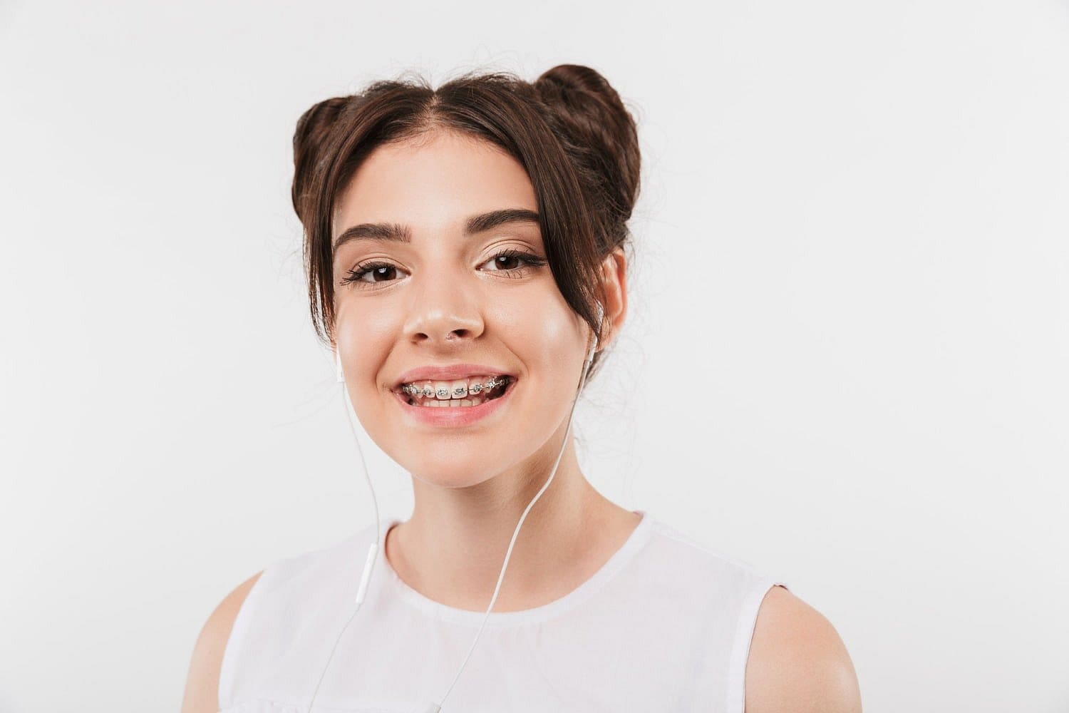 A teen girl with dark hair in two buns, wearing a white sleeveless top and listening to earphones, smiles with metal braces in this photo for Holt Orthodontics in Twinsburg, OH.