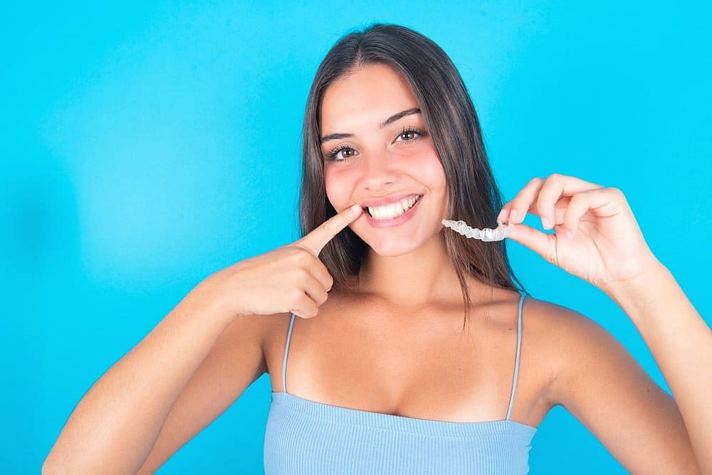 A woman in a light blue tank top smiles and points to her teeth while holding a clear aligner, highlighting that Invisalign may be covered by insurance at Holt Orthodontics in Twinsburg, OH.