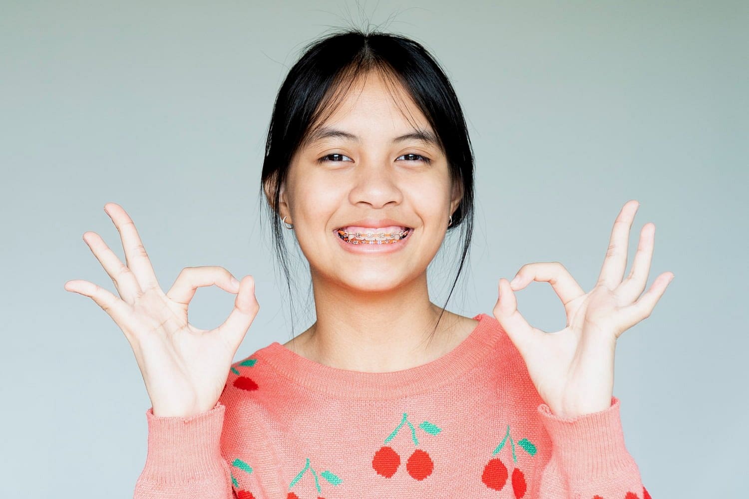 A smiling young person in a peach sweater, possibly visiting Holt Orthodontics in Twinsburg, OH for children’s orthodontic care, makes an "OK" hand gesture with both hands against a plain background.