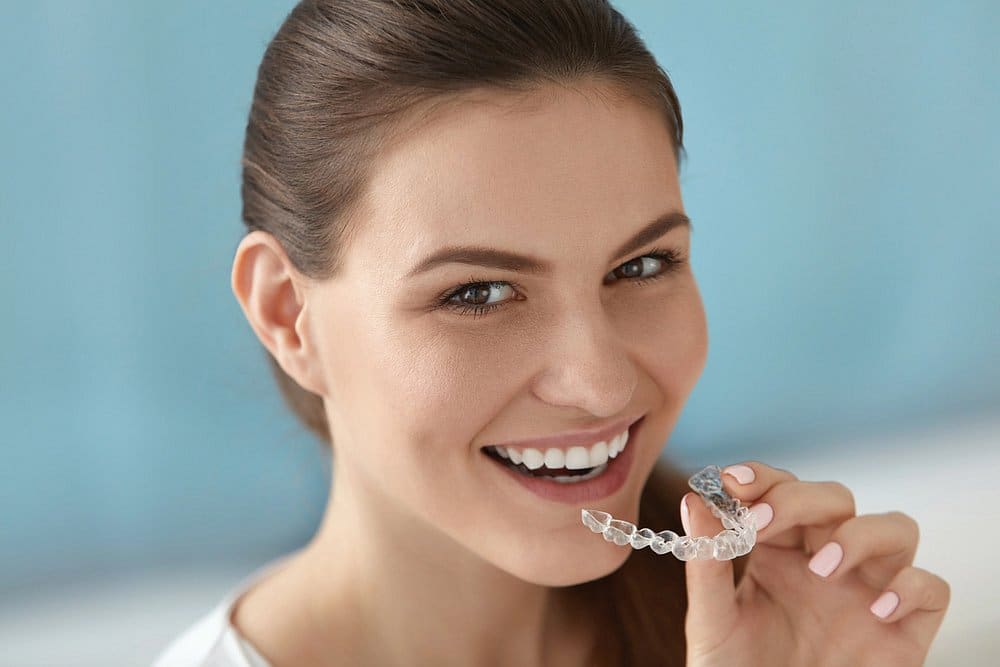 A woman smiles while holding a clear dental aligner near her mouth, highlighting the benefits of affordable Invisalign at Holt Orthodontics in Twinsburg, OH, against a light blue background.