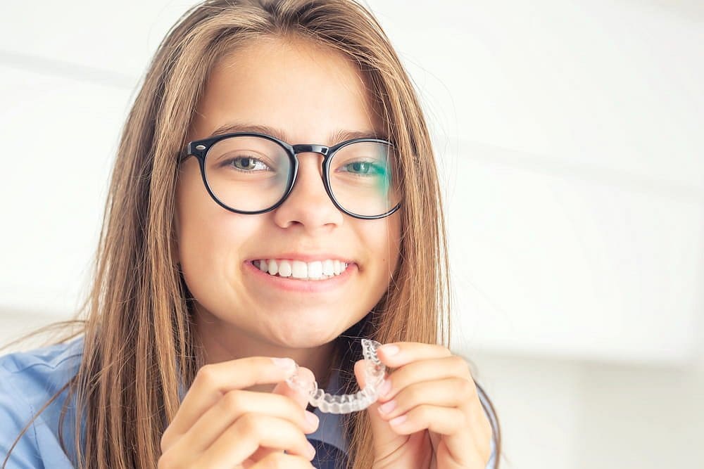 A smiling young person with glasses holds a clear aligner, showcasing the advantages of choosing Holt Orthodontics in Twinsburg, OH for children's clear aligner treatment.