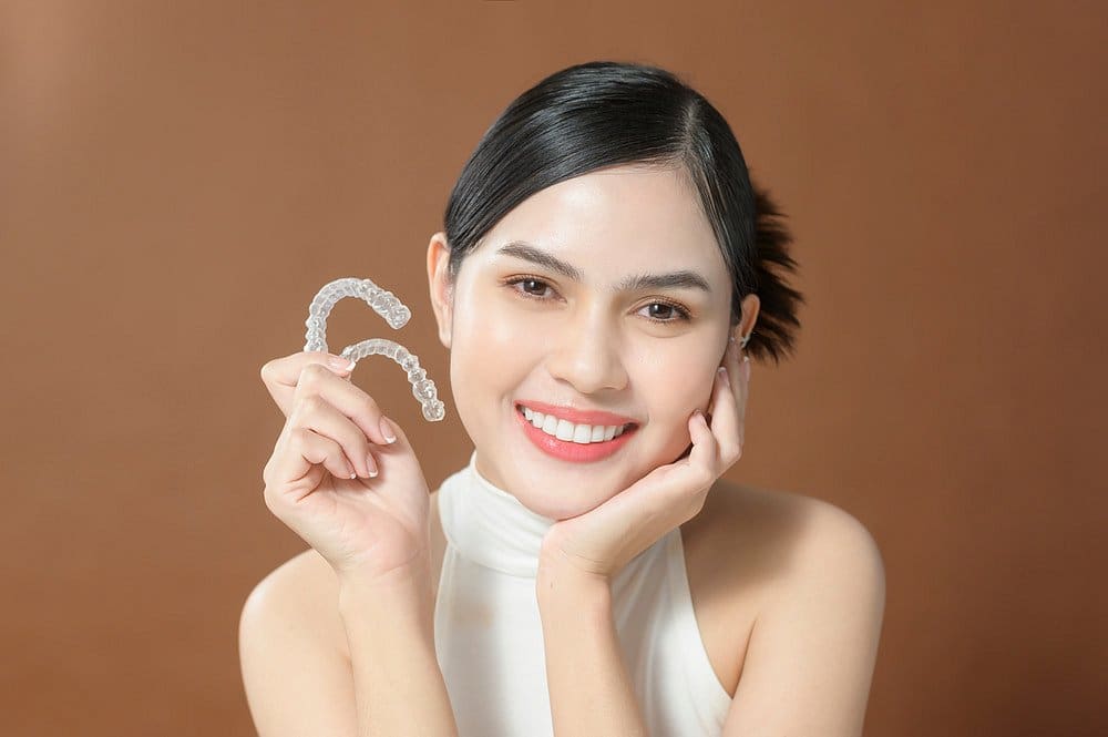 A woman smiles while holding clear dental aligners, showcasing the benefits of an Invisalign Payment Plan at Holt Orthodontics in Twinsburg, OH, against a simple brown background.