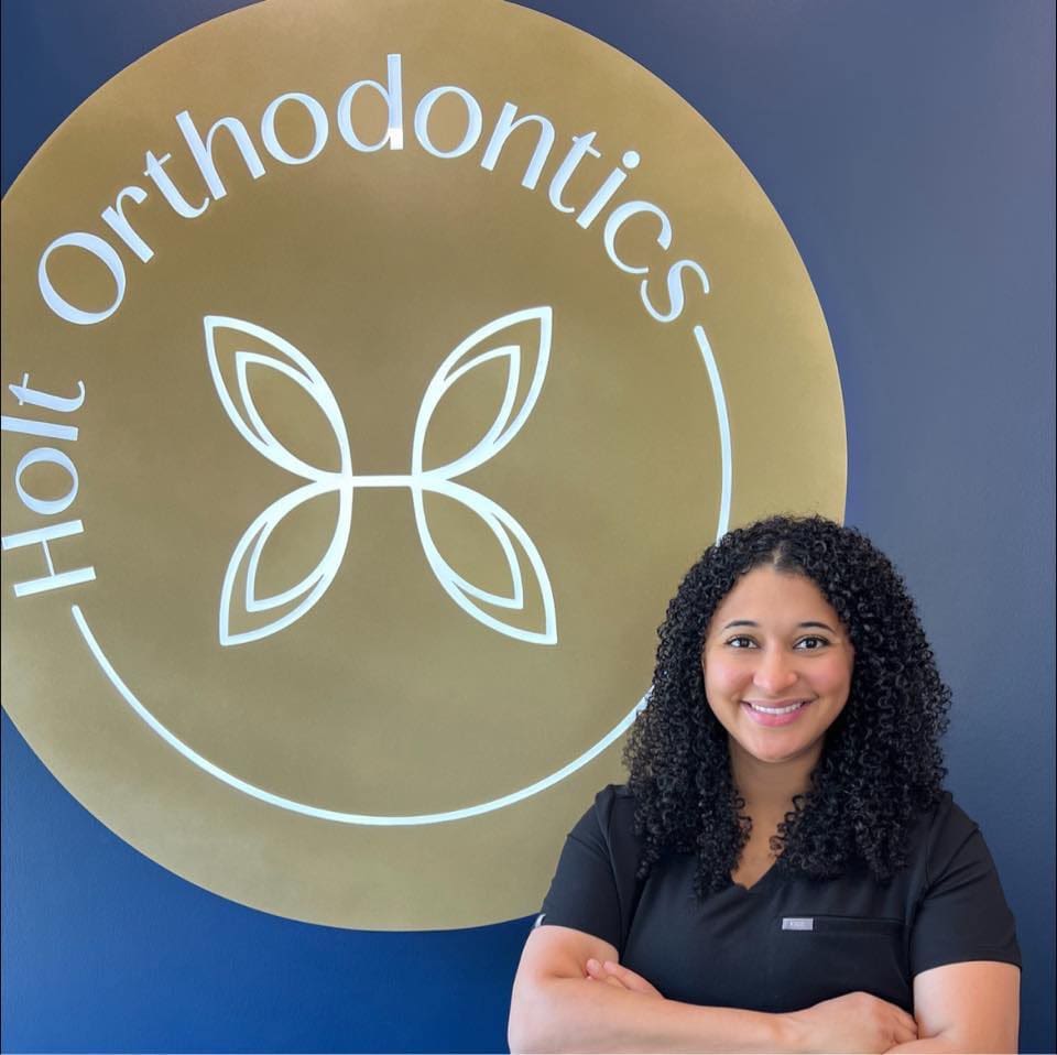 In front of a Holt Orthodontics sign on a blue and gold wall in Twinsburg, OH, Dr. Shalise Holt—wearing black scrubs and sporting curly hair—smiles warmly.