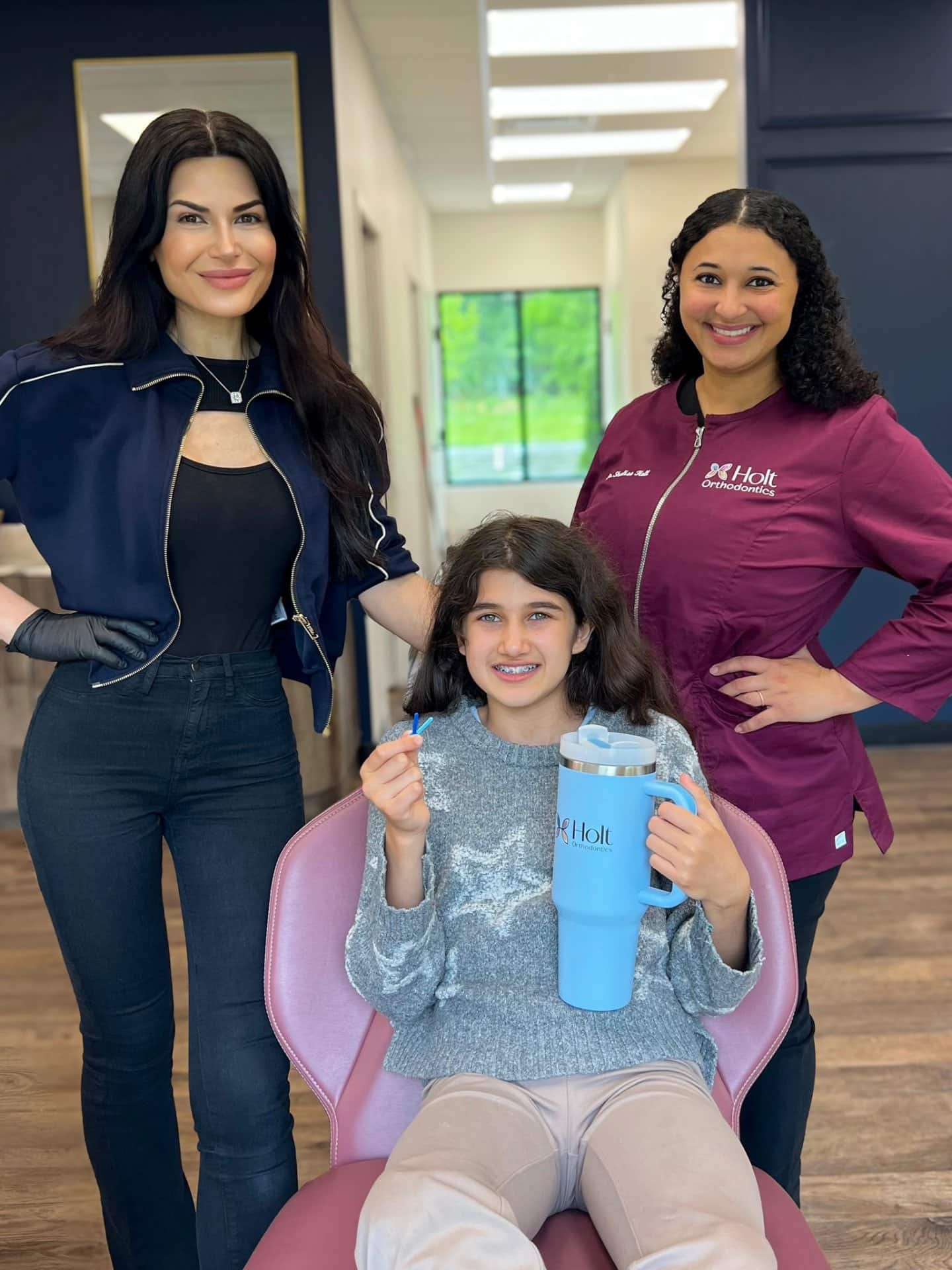 At Holt Orthodontics in Twinsburg, OH, Dr. Shalise Holt and a colleague smile in professional attire while a young girl sits in the dental chair holding a large blue cup and toothbrush.