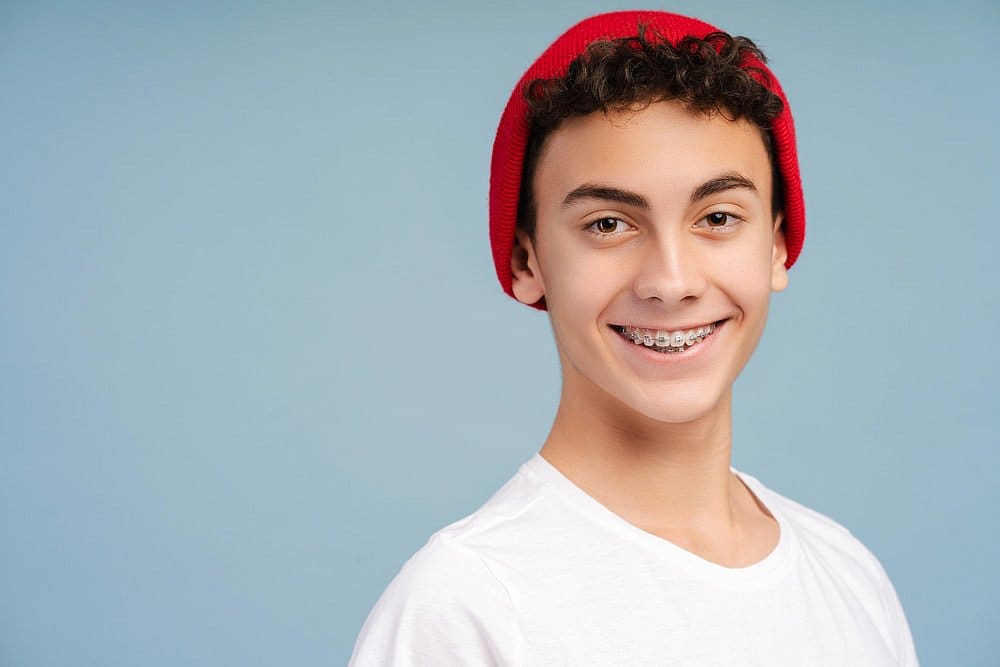 A teenager with curly hair, wearing a red beanie and white t-shirt, smiles confidently against a blue background, excited about their new smile after discovering braces cost options at Holt Orthodontics in Twinsburg, OH.