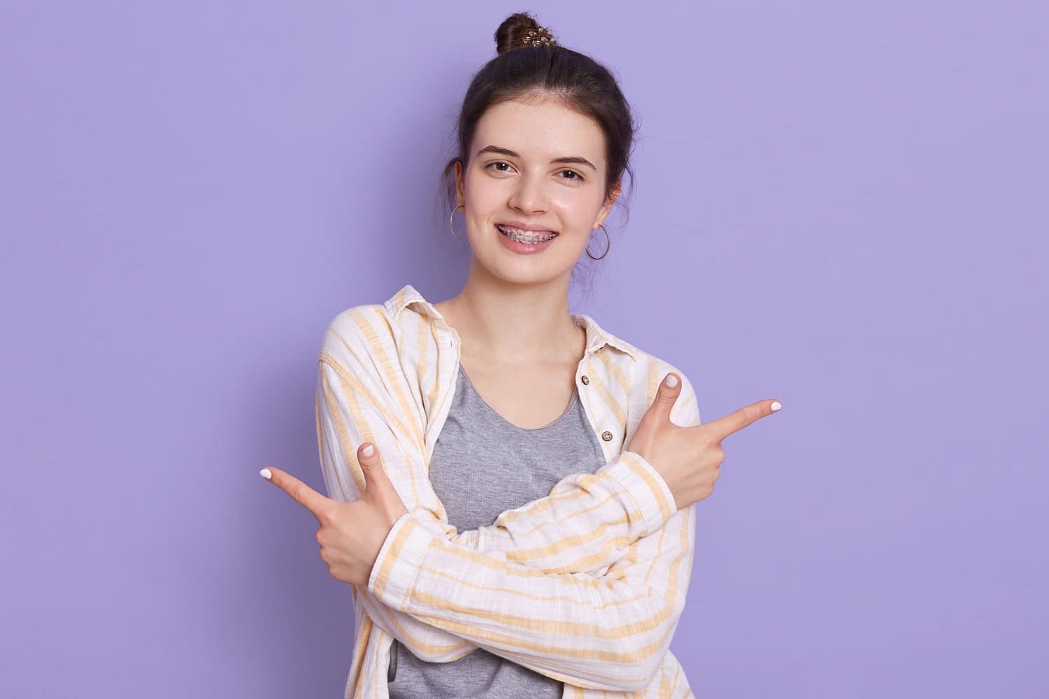 Holt Orthodontics A confident young woman with braces stands before a purple background, smiling with crossed arms and pointing both index fingers in opposite directions after discovering the affordable braces cost at Holt Orthodontics in Twinsburg, OH.