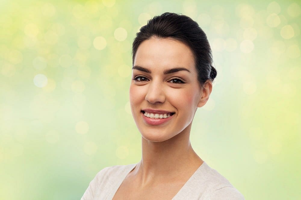 Holt Orthodontics A woman in a light-colored top smiles at the camera, showing off her clear braces from Holt Orthodontics in Twinsburg, OH, against a softly blurred green and yellow background.