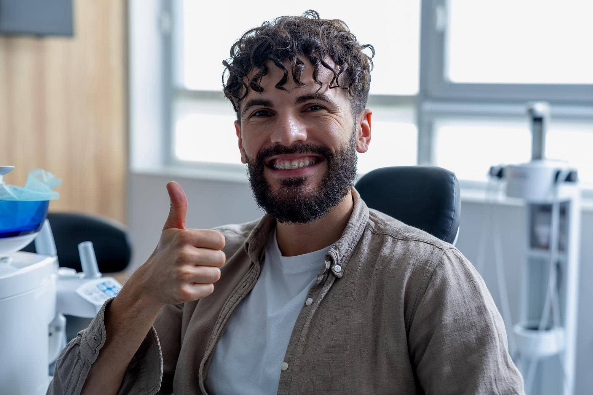 A man with curly hair and a beard sits indoors at Holt Orthodontics in Twinsburg, OH, smiling and giving a thumbs-up as he proudly shows off his clear braces.