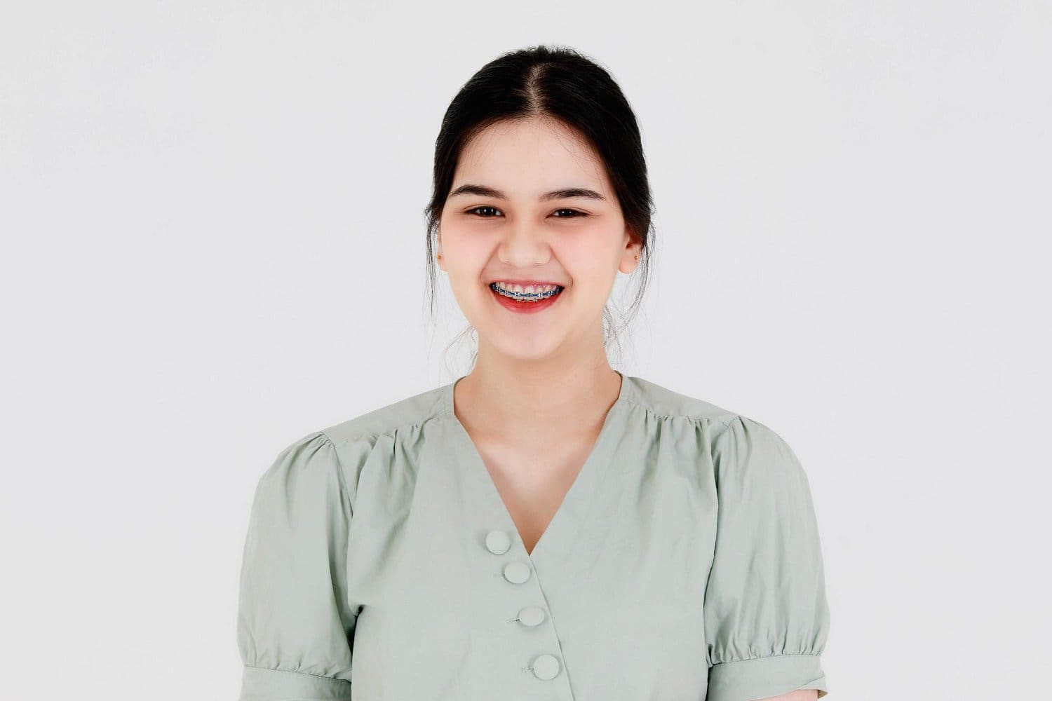 Holt Orthodontics A young woman with dark hair in a light green blouse smiles confidently at the camera, proudly displaying her braces—a reflection of the investment in her smile—against a plain white background at Holt Orthodontics in Twinsburg, OH.