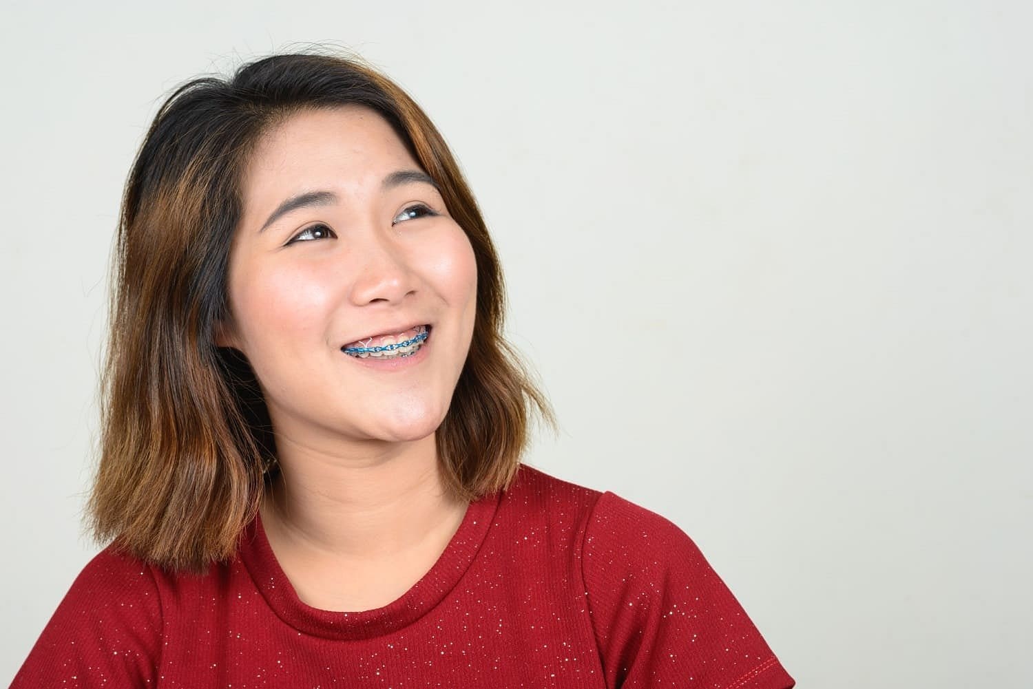 A young woman with shoulder-length brown hair and a red shirt smiles brightly, revealing blue adult braces, at Holt Orthodontics in Twinsburg, OH.
