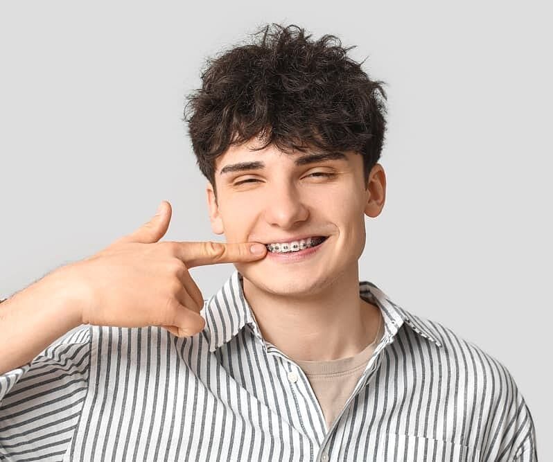 A young man in a striped shirt smiles and points to his metal braces in a photo for Holt Orthodontics in Twinsburg, OH, set against a plain gray background.