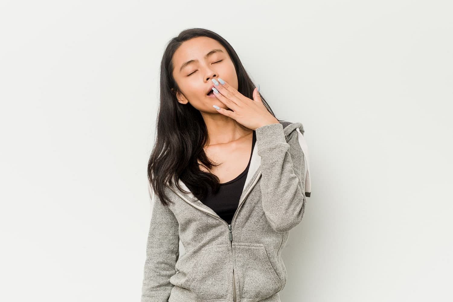 At Holt Orthodontics in Twinsburg, OH, a woman in a gray hoodie stands against a plain white background with her eyes closed, covering her mouth as she represent Causes and Risk Factors underbite.