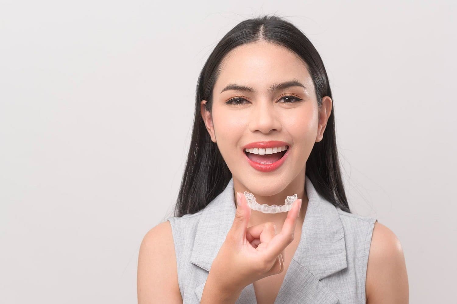 A woman with long dark hair smiles and holds a clear dental aligner in front of her mouth, highlighting how Invisalign may be covered by insurance at Holt Orthodontics in Twinsburg, OH.