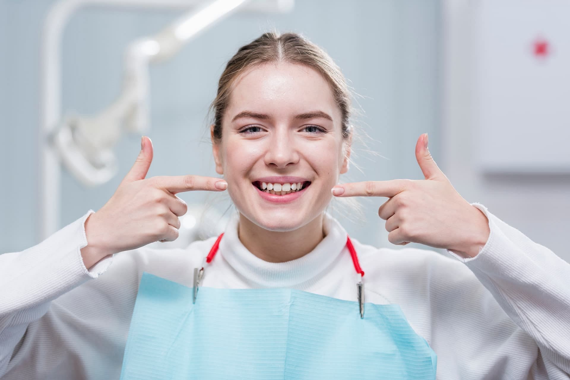 A young woman at Holt Orthodontics in Twinsburg, OH smiles while pointing to a gap between her front teeth, wearing a white top and blue dental bib, possibly discussing her overbite.