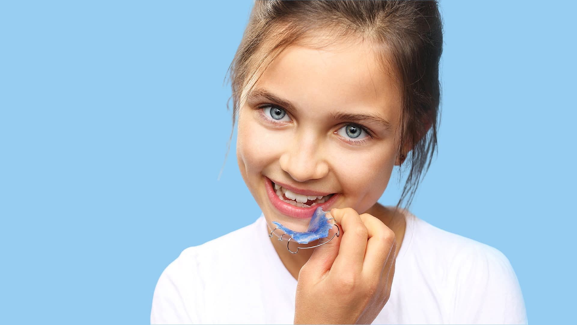 A young girl with blue eyes smiles while holding an orthodontic appliance near her mouth against a light blue background at Holt Orthodontics in Twinsburg, OH.