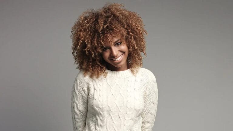A person with curly hair and a white cable-knit sweater smiles subtly against a plain gray background at Holt Orthodontics in Twinsburg, OH.