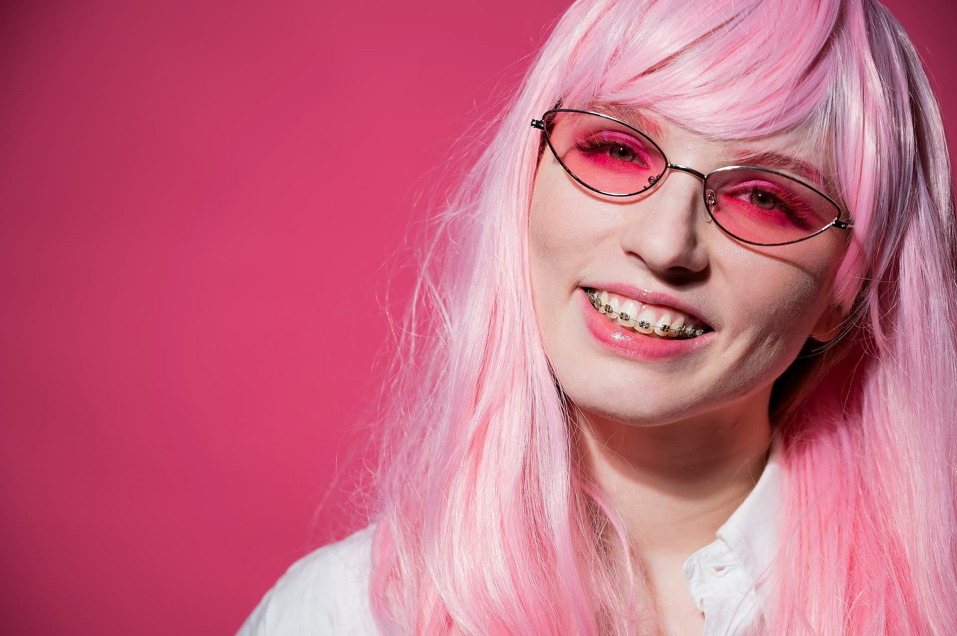Smiling at the camera against a solid pink background, a woman with long pink hair, pink-tinted glasses, and gold braces showcases their style—reflecting the vibrant results possible at Holt Orthodontics in Twinsburg, OH.