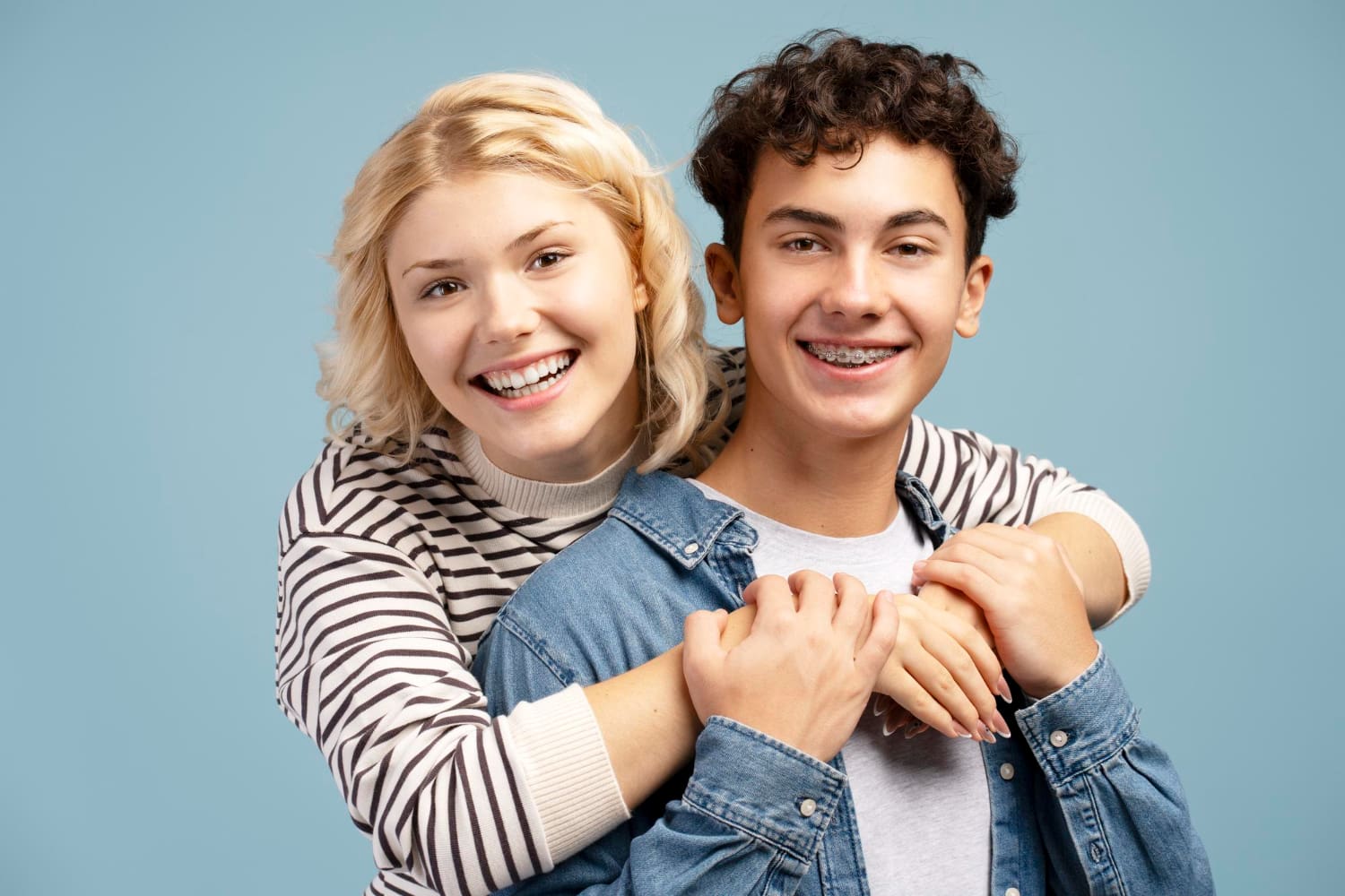 Smiling teens, one with blonde hair and one with braces, hug in front of a blue background at Holt Orthodontics in Twinsburg, OH.
